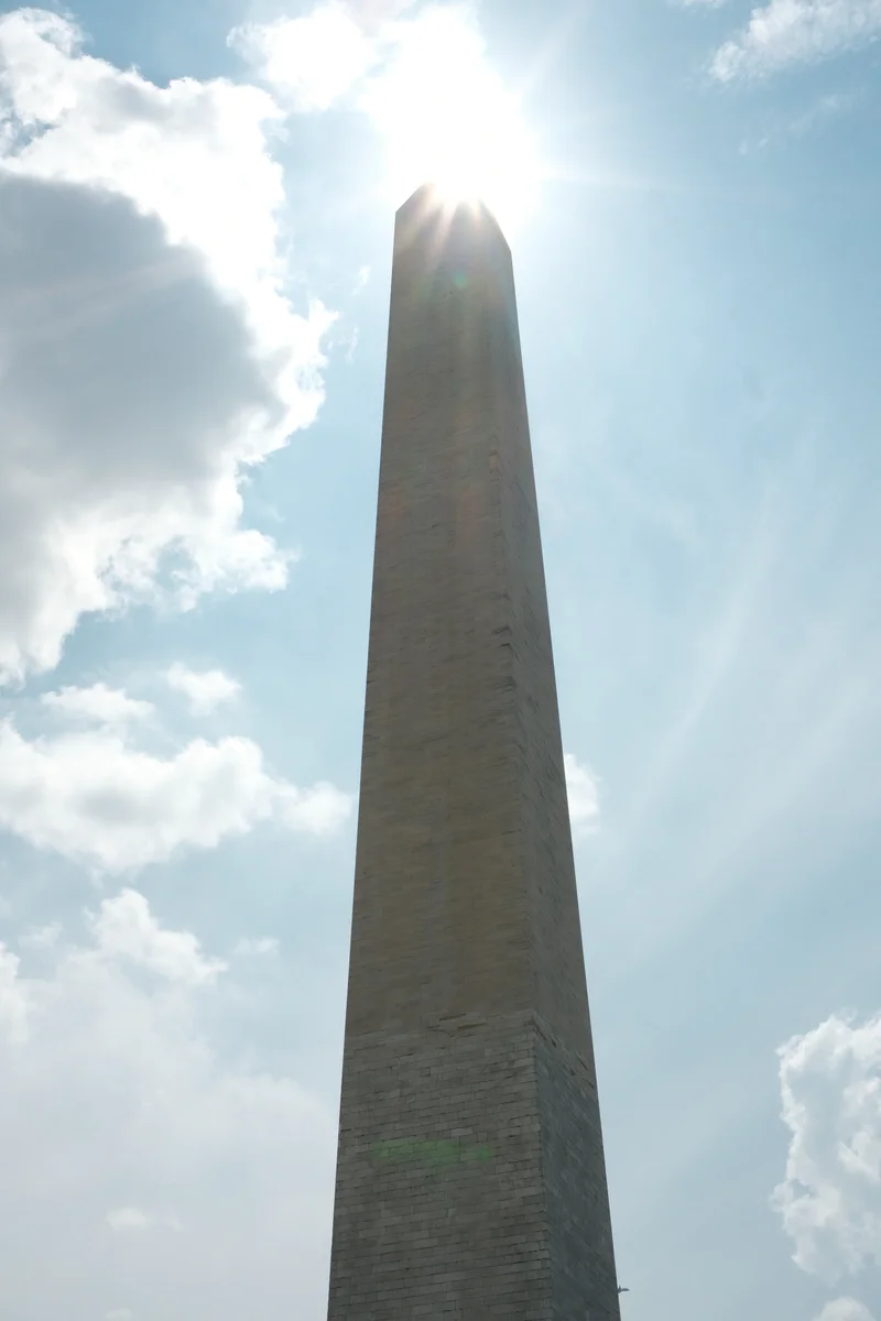 Sunlight breaks around the tip of the Washington Monument beneath a blue, cloud-streaked sky.