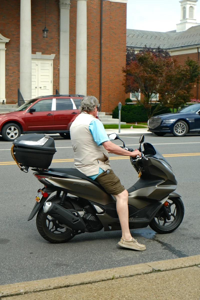 A rider waits on a scooter at a street corner in front of a red-brick building.