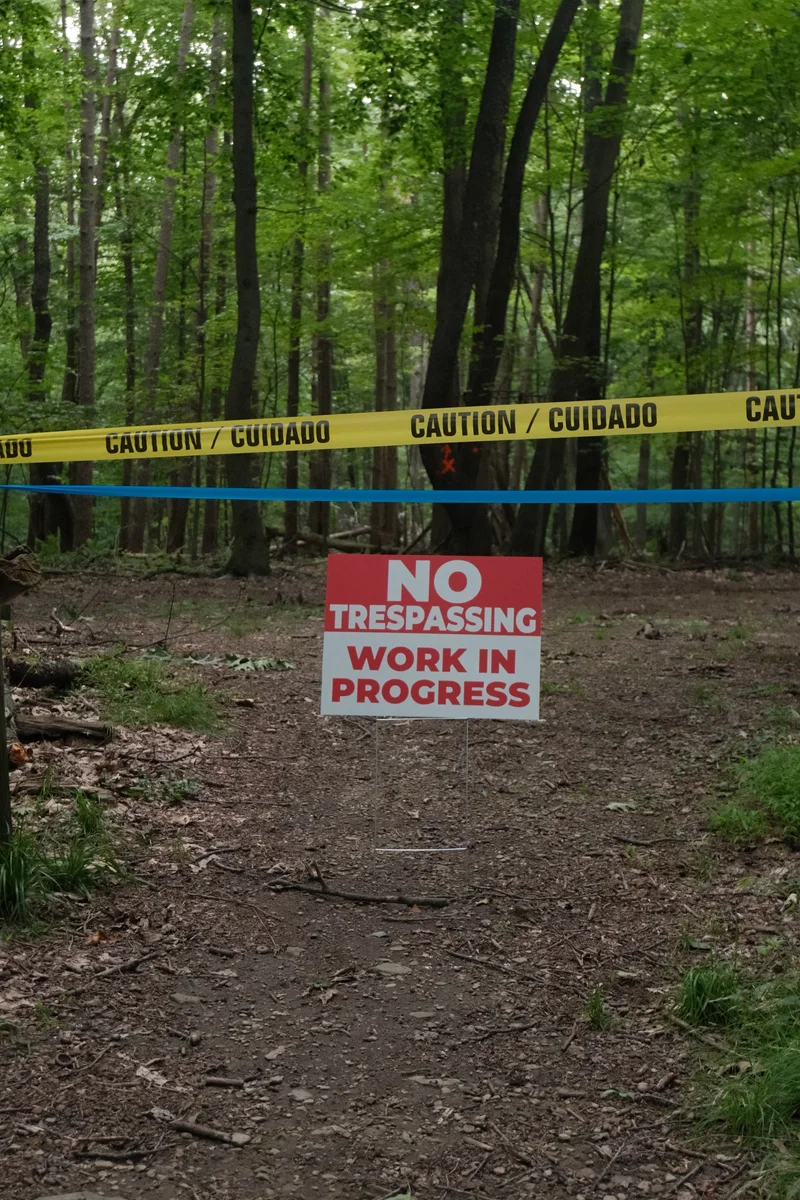 Caution tape and a no-trespassing sign interrupt a trail beneath a canopy of green trees.