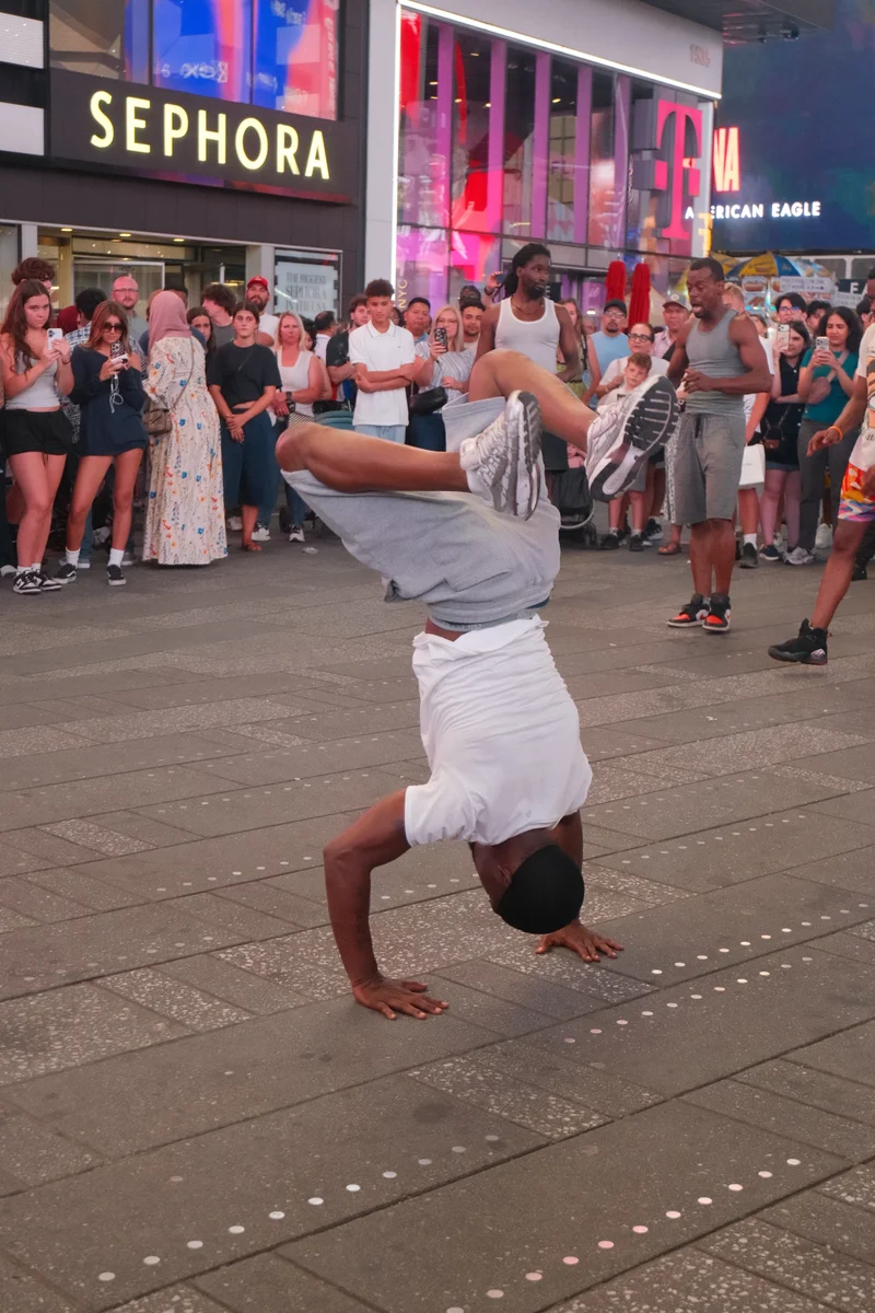 A street performer balances upside down while a crowd watches beneath the bright signs of Times Square.
