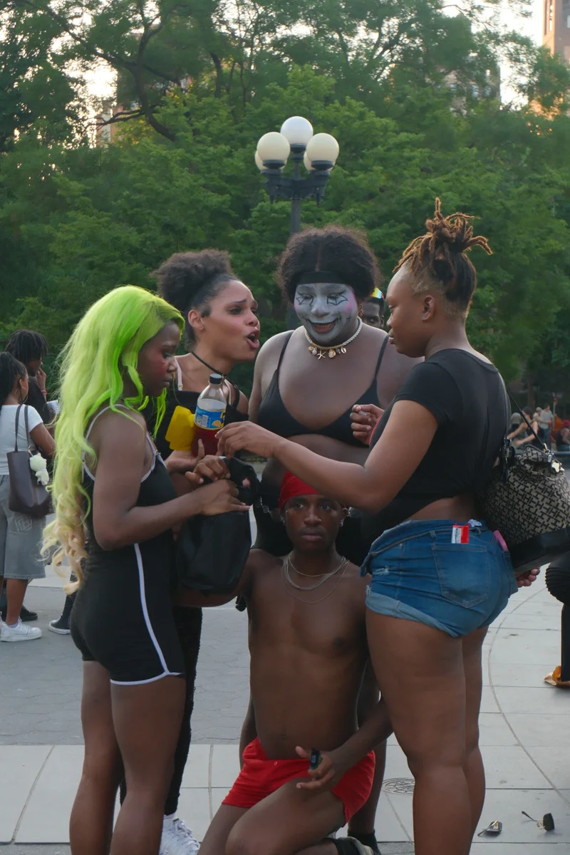 People gather around a performer with a painted face, surrounded by trees and the energy of a public square.