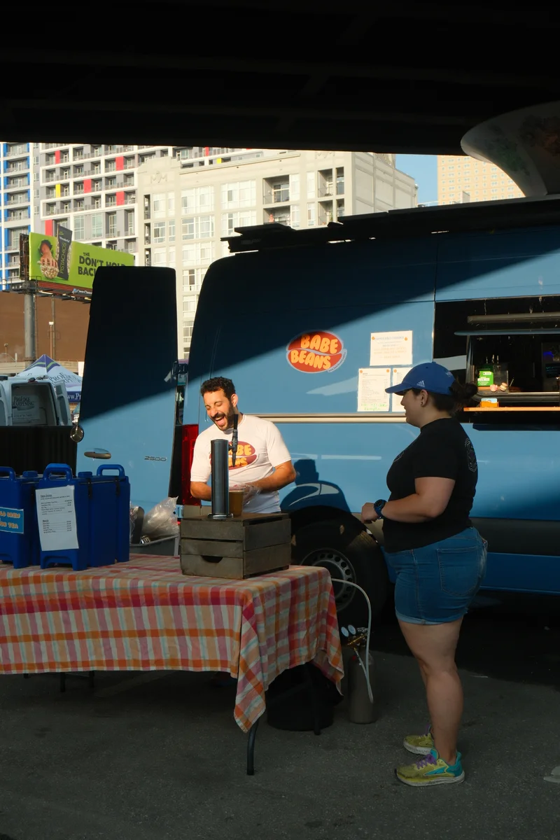 Two people stand beside a checkered market table in the shade of an overpass.