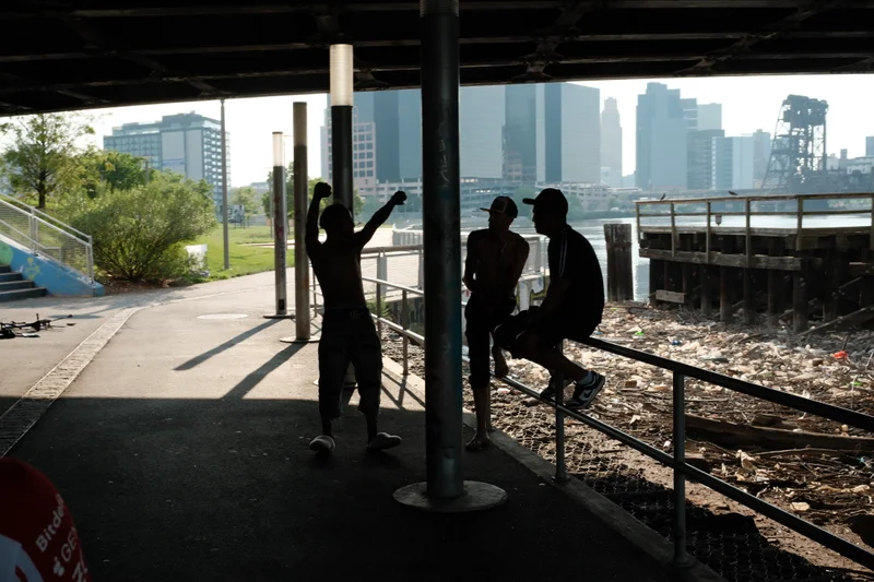 Silhouettes pause beneath a waterfront overhang, framed by columns and the distant city skyline.